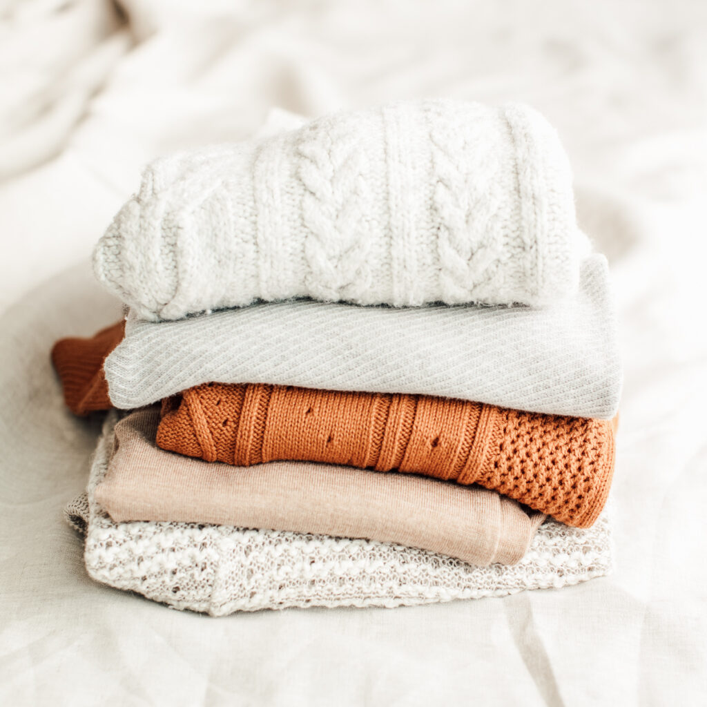 A pile of clothing from future blog collaborations, a white knitted jumper folded ontop of a grey jumper, ontop of an orange jumper